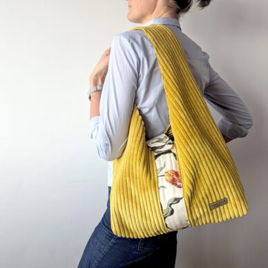 Kobieta prezentuje dużą, musztardową torbę na ramię wykonaną z grubego sztruksu tapicerskiego. Torba ma fason typu hobo, posiada szeroki pas na ramię oraz pionowy ozdobny panel z jasnego materiału w kolorowe tulipany. Widoczna jest również mała, beżowa metka z napisem "Lidia Pilch Handmade".